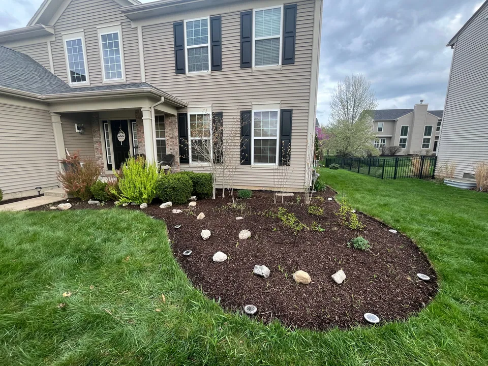 Mulched planting bed with shrubs and stones beside a tan house