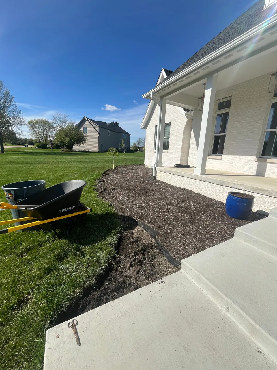 Mulched front bed with a wheelbarrow and tools beside a white porch